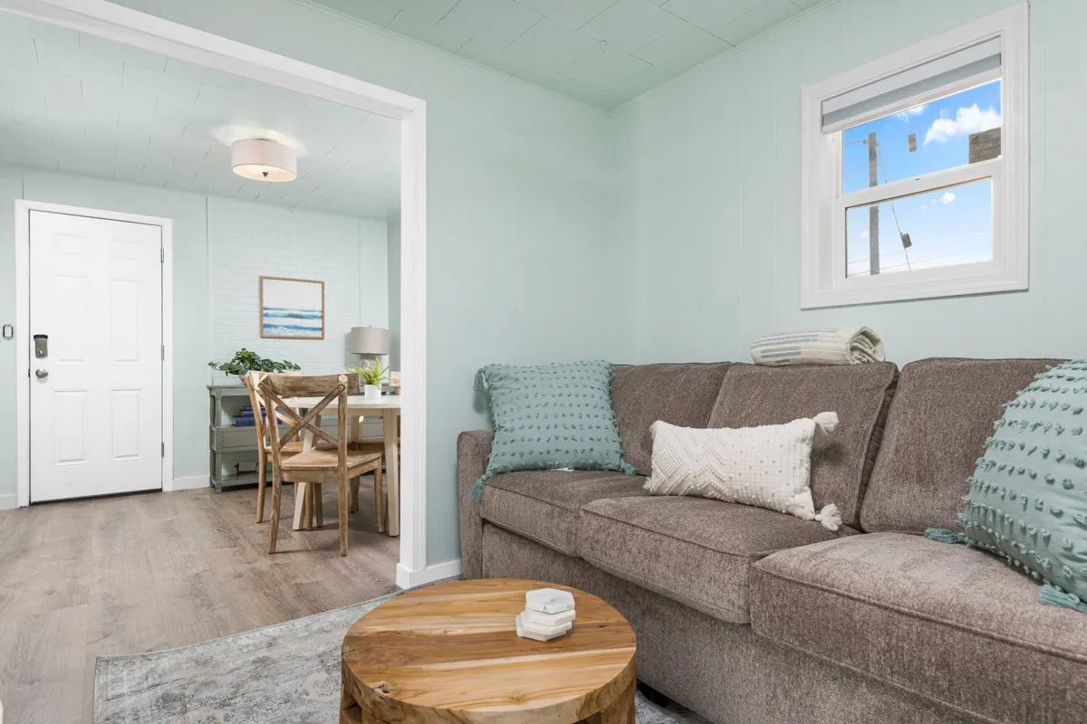 The image features a cozy, inviting living space with a plush sofa, stylish wooden coffee table, vibrant artwork on the wall, and a glimpse of the adjoining dining area through a doorway. The soft lighting and thoughtful decor create a warm, welcoming atmosphere ideal for relaxing or gathering with loved ones.