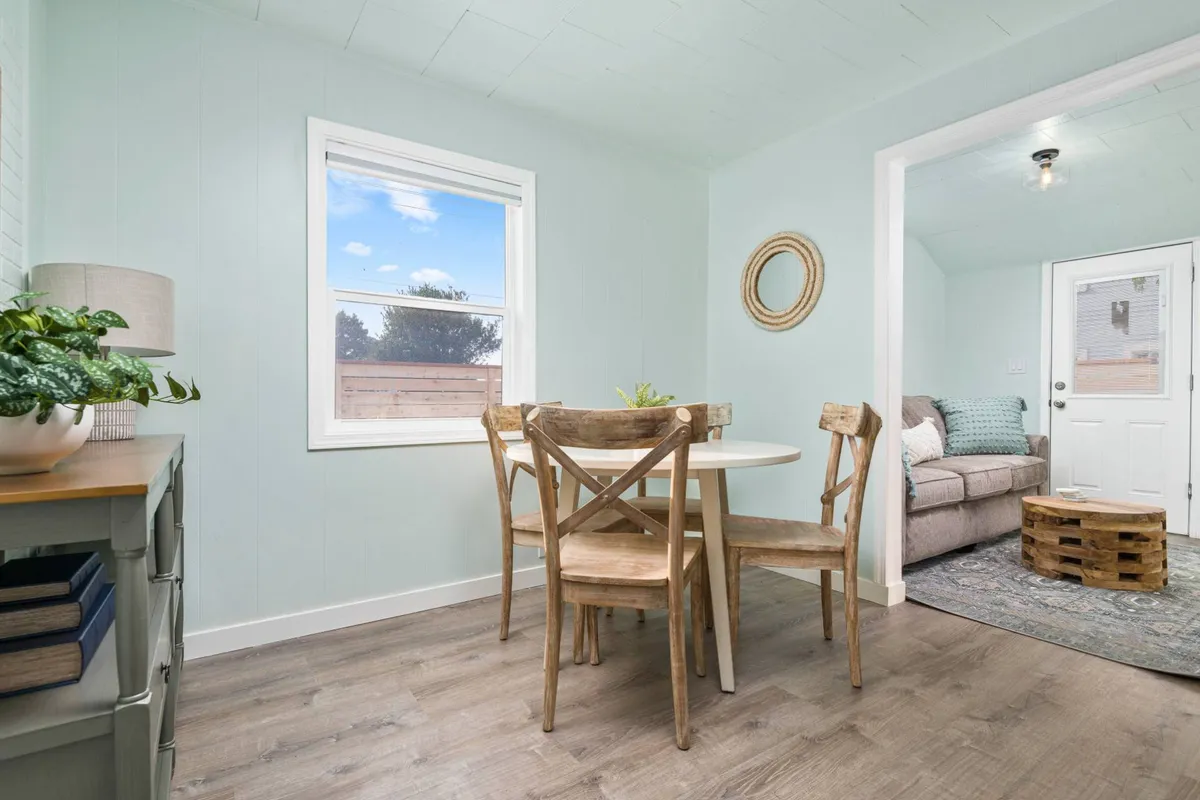 This image showcases the cottage's front door, visible from a welcoming dining and living area with a table, chairs, sofa, and a decorative wall mirror. The space exudes an inviting atmosphere that suggests both comfort and the ease of coming and going for day trips.