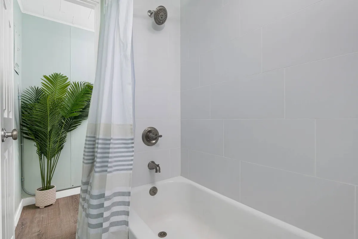 A serene bathroom corner featuring a white bathtub with a stylish shower fixture and a lush potted plant, set against a calming backdrop. Soft lighting and a simple shower curtain evoke a tranquil, spa-like atmosphere.