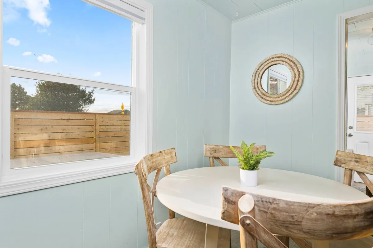 A cozy dining area with a round table, wooden chairs, a large window flooding the space with natural light, and stylish decor including a wall mirror and indoor plant. The setting is perfect for casual dining, morning coffee, or family celebrations.