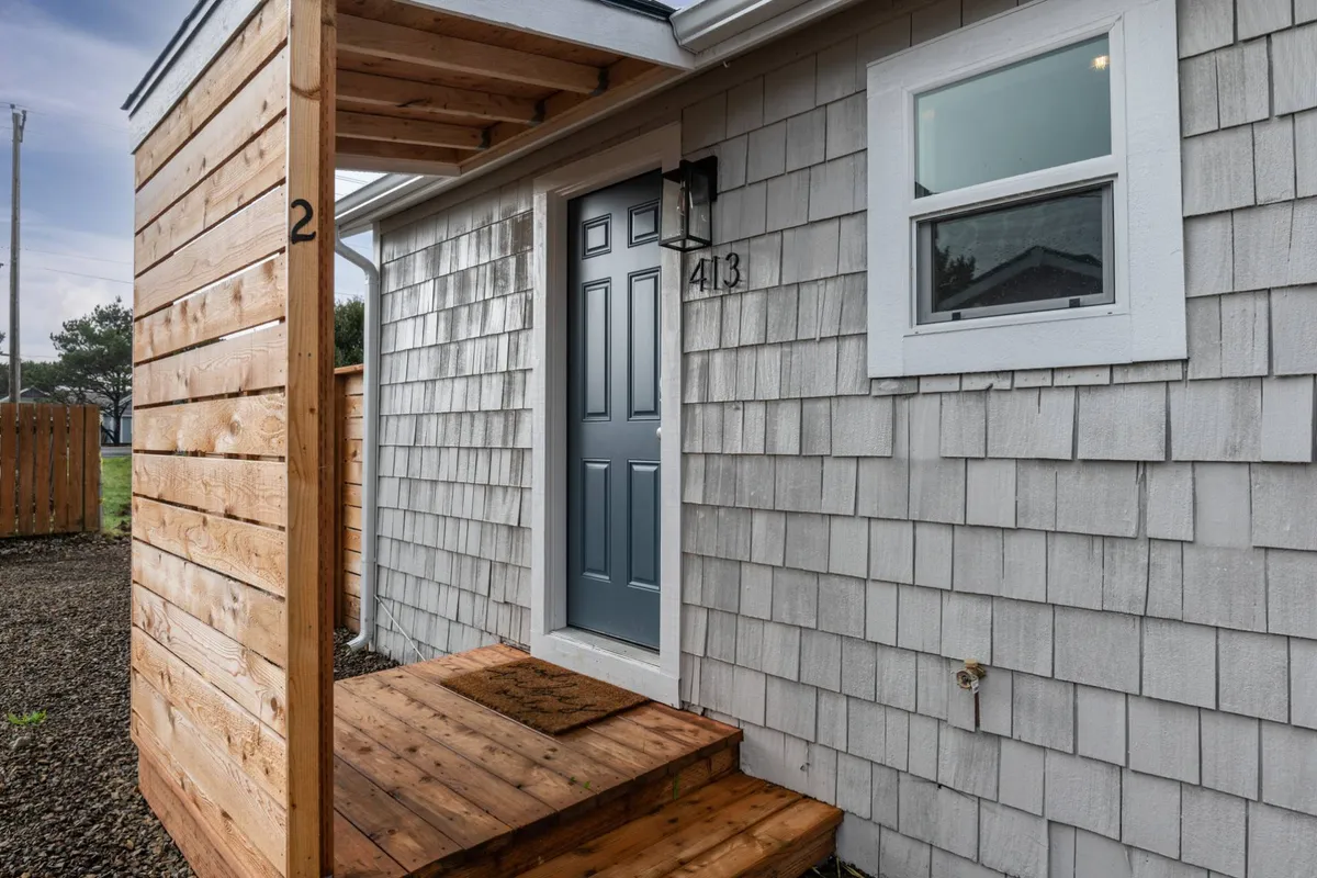 This image captures the exterior entrance of the cottage, featuring an inviting front door, wooden steps, and classic shingle siding. The outdoor setting hints at the private amenities available just beyond the entrance, all while conveying a warm and welcoming mood.