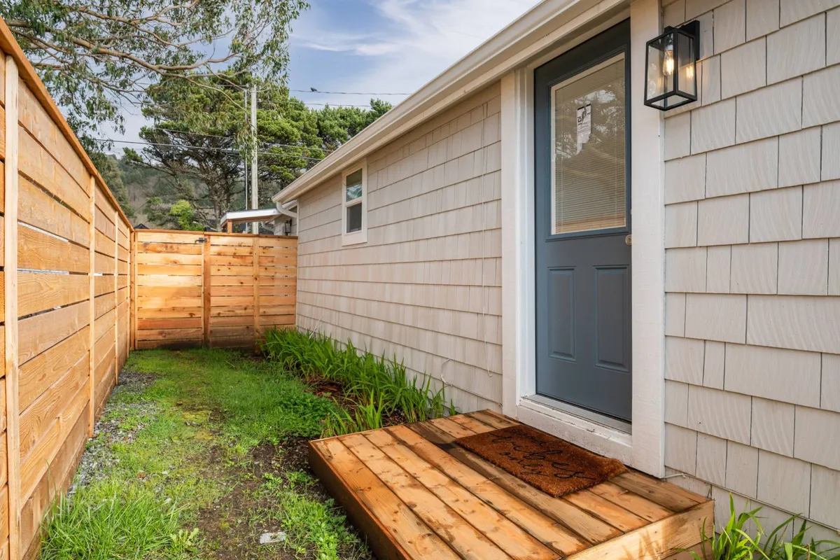 This image shows the front yard and entryway of the cottage, with a focus on a welcoming front door, wooden steps, and a garden area bordered by a visible fence. The inviting outdoor space suggests a comfortable and safe spot for guests and their pets to enjoy time outdoors.