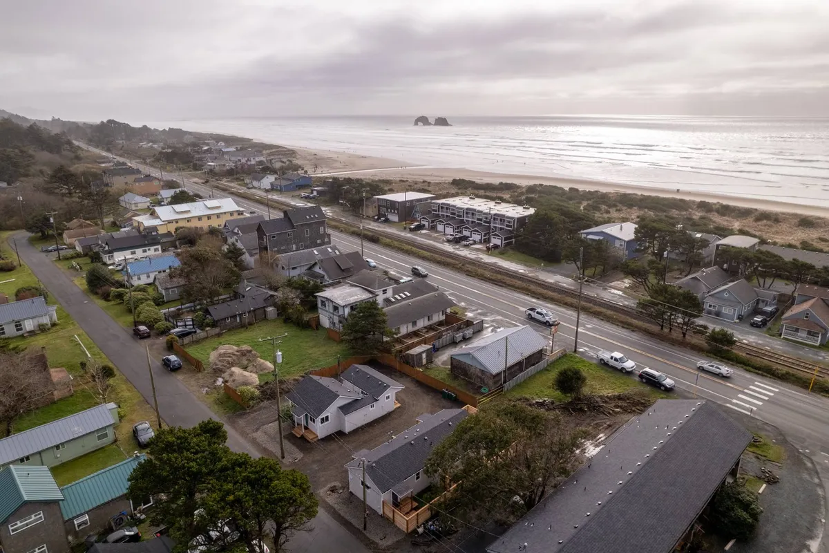 This image presents a sweeping view of Rockaway Beach, featuring charming coastal houses, a walkable road, the expansive ocean, and natural sand dunes. The scene captures a tranquil atmosphere, inviting viewers to imagine strolling the streets, visiting local shops and eateries, and soaking in the surrounding beauty.