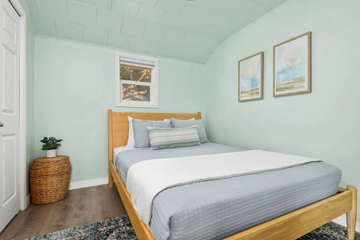 A tranquil bedroom featuring a full-size bed dressed in soft linens, flanked by a bedside table and lamp. Decorative wall paintings and a lush green plant add to the soothing atmosphere. Soft morning light filters through a window, illuminating a woven basket at the foot of the bed, reinforcing the room’s cozy, restful ambiance.
